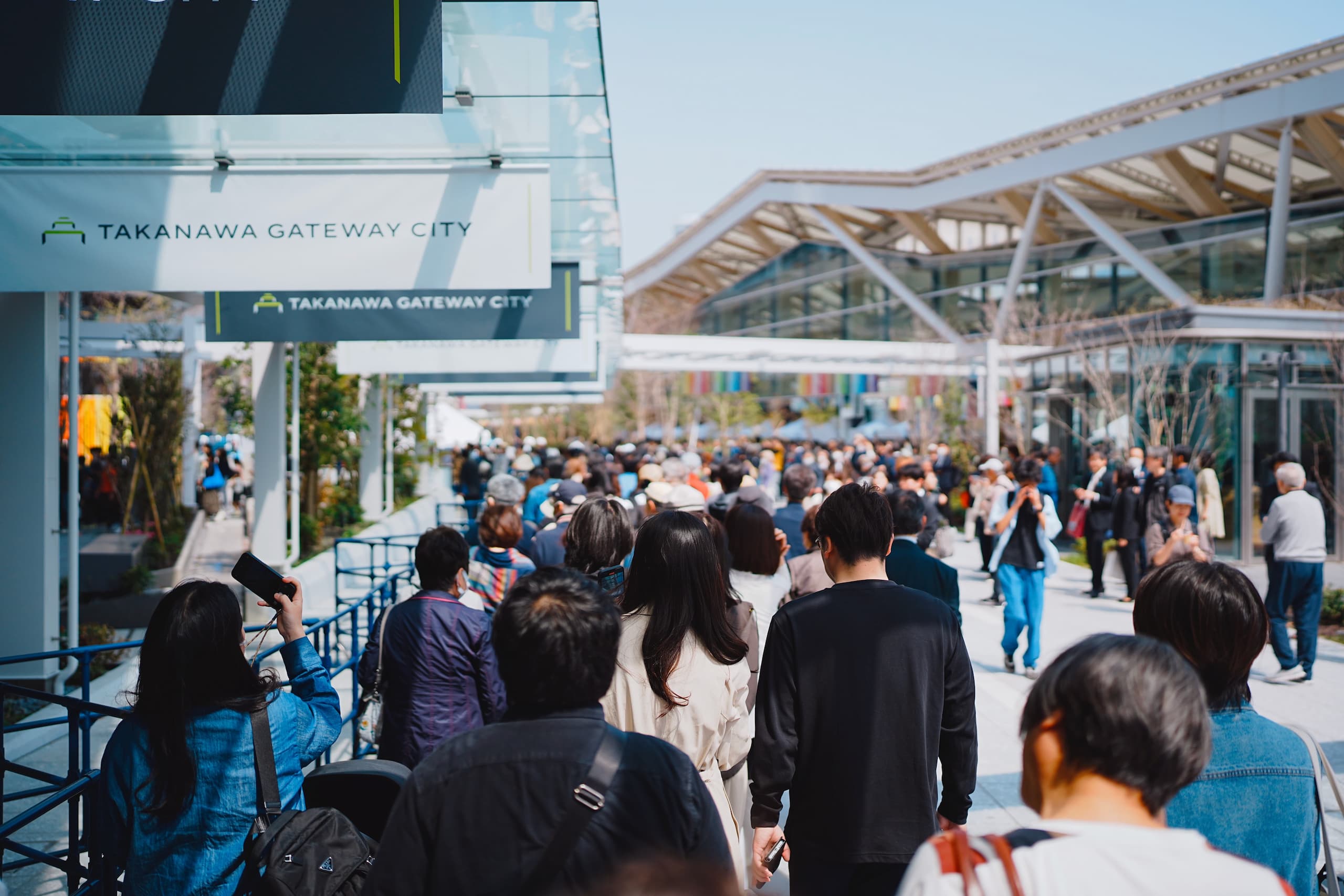 多くの人々が高輪ゲートウェイ駅付近の屋外の広い歩道を歩いている様子。